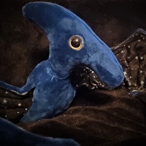May include: A plush toy in the shape of a blue sea creature with a large, round eye. The toy has a tail and fins, and is made of soft, velvety material. The background is a dark, textured fabric.