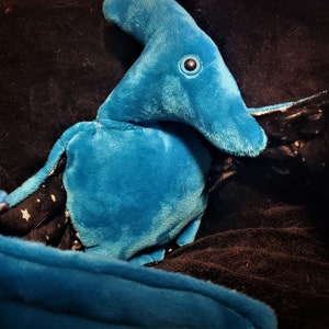 May include: A plush blue pterodactyl toy with a long beak and large eye. The wings are black with a white star pattern. The toy is made of soft material and is lying on a dark surface.