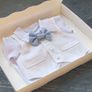 K&ouml;nnte beinhalten: Ein wei&szlig;es Baby-Outfit mit Weste und einer blauen Fliege. Das Outfit befindet sich in einer wei&szlig;en Box.