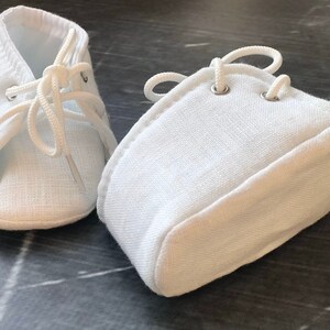 K&ouml;nnte beinhalten: Ein Paar wei&szlig;e Babyschuhe mit Schn&uuml;rsenkeln. Die Schuhe sind aus weichem, wei&szlig;em Stoff und haben ein schlichtes Design.