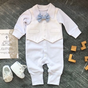 K&ouml;nnte beinhalten: Wei&szlig;er Baby-Strampler mit einer wei&szlig;en Weste und einer hellblauen Fliege. Der Strampler hat einen Knopfverschluss am Schritt. Das Baby tr&auml;gt wei&szlig;e Schuhe. Das Bild zeigt auch eine wei&szlig;e Karte mit schwarzem Text und einer goldenen Spitzen&uuml;berlagerung.