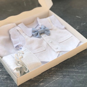 K&ouml;nnte beinhalten: Ein wei&szlig;es Baby-Outfit-Set in einer wei&szlig;en Box. Das Set beinhaltet eine wei&szlig;e M&uuml;tze, eine wei&szlig;e Weste mit einer blauen Schleife, einen wei&szlig;en Strampler und wei&szlig;e Schuhe.