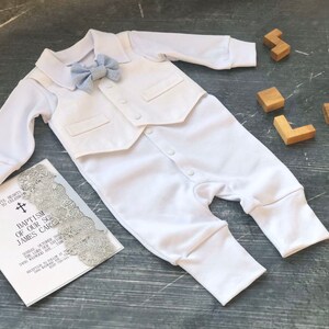 K&ouml;nnte beinhalten: Wei&szlig;es Baby-Outfit mit Weste und Fliege. Das Outfit liegt auf einer grauen Oberfl&auml;che mit einer wei&szlig;en Einladungskarte, auf der "Taufe unseres Sohnes James Carl" steht.