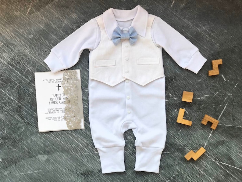K&ouml;nnte beinhalten: Wei&szlig;er Baby-Strampler mit einer wei&szlig;en Weste und einer hellblauen Fliege. Der Strampler hat einen Kragen und lange &Auml;rmel. Die Weste hat zwei Taschen. Die Fliege ist um den Hals gebunden.