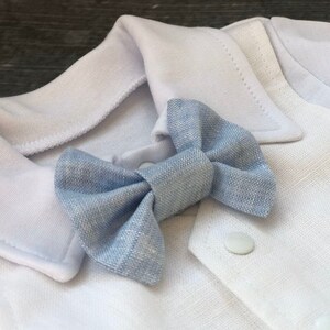 K&ouml;nnte beinhalten: Ein wei&szlig;es Baby-Outfit mit einer hellblauen Fliege. Das Outfit hat einen Kragen und einen Knopfverschluss.