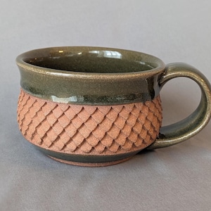 May include: Handmade ceramic mug with a textured, scale-like pattern in a warm terracotta color. The rim and handle are a deep olive green. This unique mug is perfect for enjoying coffee, tea, or other beverages.