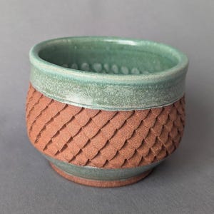 May include: Handmade ceramic bowl with a textured, scale-like pattern in a reddish-brown color. The rim and base are a muted green, creating a two-tone effect. The bowl is designed for serving or display.