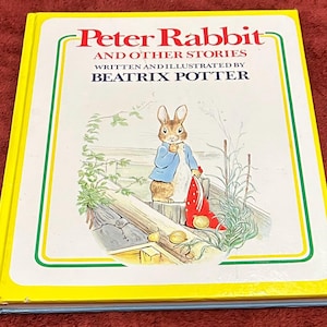 May include: A hardcover book titled Peter Rabbit and Other Stories written and illustrated by Beatrix Potter. The white cover features a yellow border and a central illustration of a brown rabbit wearing a blue jacket, holding a red cloth.
