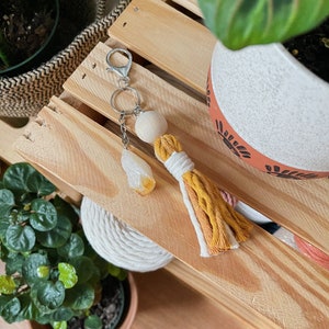 Puede incluir: Un llavero de cuentas de madera con un borla blanco y amarillo. La borla está hecha de hilo y tiene una cuenta de madera natural en la parte superior. El llavero está colgado en una estantería de madera.