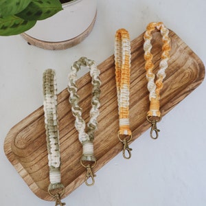Op de afbeelding: Drie sleutelhangers met verschillende kleurcombinaties van wit en geel, wit en groen en wit en oranje. De sleutelhangers zijn gemaakt van gevlochten garen en hebben een gouden metalen sluiting.