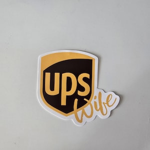 May include: A sticker featuring the UPS logo in brown and gold, with the text "UPS Wife" in a cursive font. The sticker has a white border and is on a white background.