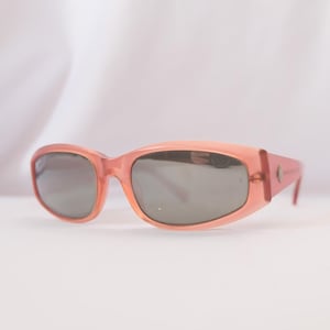 Pode incluir: &Oacute;culos de sol cor de p&ecirc;ssego com lentes cinza escuro. Os &oacute;culos t&ecirc;m uma arma&ccedil;&atilde;o retangular arredondada e um pequeno emblema nas hastes. As hastes s&atilde;o ligeiramente curvas e as lentes s&atilde;o tingidas.
