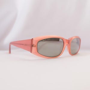 Pode incluir: &Oacute;culos de sol cor de p&ecirc;ssego com lentes cinzentas. As arma&ccedil;&otilde;es retangulares t&ecirc;m uma forma ligeiramente arredondada e hastes retas. Os &oacute;culos de sol est&atilde;o sobre um fundo branco.