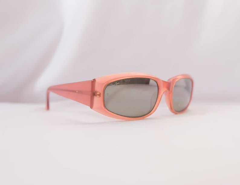Pode incluir: &Oacute;culos de sol cor de p&ecirc;ssego com lentes cinzentas. As arma&ccedil;&otilde;es retangulares t&ecirc;m uma forma ligeiramente arredondada e hastes retas. Os &oacute;culos de sol est&atilde;o sobre um fundo branco.