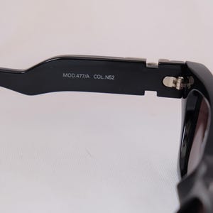 以下が含まれることがあります： 太いフレームの黒いサングラスのクローズアップ。テンプルには「MOD.477/A COL.N52」の文字がプリントされています。レンズは濃い色です。ヒンジはシルバーです。