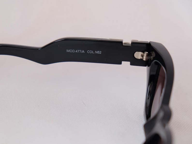 以下が含まれることがあります： 太いフレームの黒いサングラスのクローズアップ。テンプルには「MOD.477/A COL.N52」の文字がプリントされています。レンズは濃い色です。ヒンジはシルバーです。
