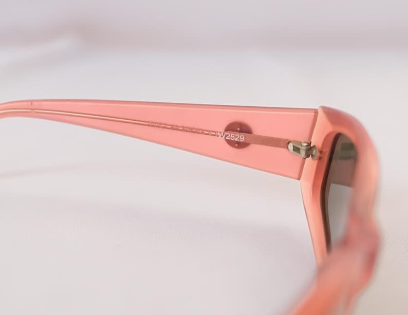 Pode incluir: Close-up de &oacute;culos de sol cor-de-rosa. A arma&ccedil;&atilde;o &eacute; de cor s&oacute;lida e transl&uacute;cida. A dobradi&ccedil;a e a haste s&atilde;o vis&iacute;veis, com o texto "W2529" impresso na haste. As lentes s&atilde;o de cor escura.