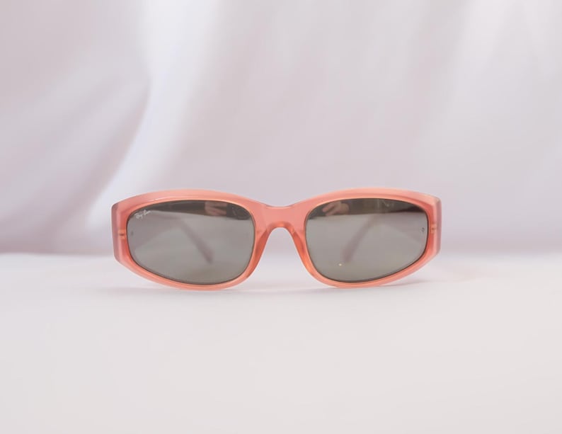 Pode incluir: Um par de &oacute;culos de sol cor de coral com lentes cinzentas. Os &oacute;culos t&ecirc;m uma arma&ccedil;&atilde;o retangular e um log&oacute;tipo subtil na lente esquerda. O fundo &eacute; branco suave.