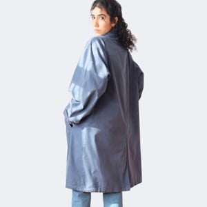May include: A long, slate blue coat with a collar and pockets. The coat is knee-length and appears to be made of a smooth, lightweight fabric. The person is wearing blue jeans. The coat is a versatile outerwear piece.