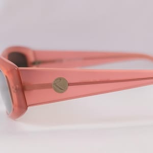 Pode incluir: Close-up de &oacute;culos de sol cor de coral com lentes escuras. As arma&ccedil;&otilde;es t&ecirc;m formato retangular com bordas arredondadas. Um pequeno emblema circular est&aacute; na haste. Os &oacute;culos de sol est&atilde;o em um fundo branco suave.