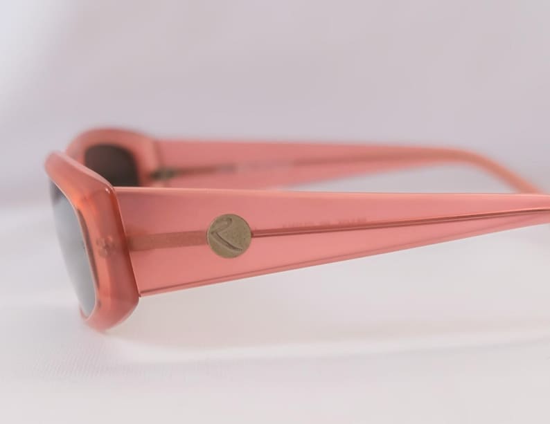 Pode incluir: Close-up de &oacute;culos de sol cor de coral com lentes escuras. As arma&ccedil;&otilde;es t&ecirc;m formato retangular com bordas arredondadas. Um pequeno emblema circular est&aacute; na haste. Os &oacute;culos de sol est&atilde;o em um fundo branco suave.