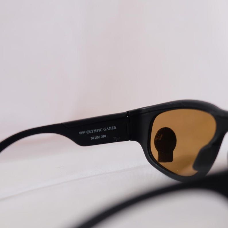 May include: Black sunglasses with brown lenses. The sunglasses have "1992 Olympic Games 36 USC 380" printed on the side.