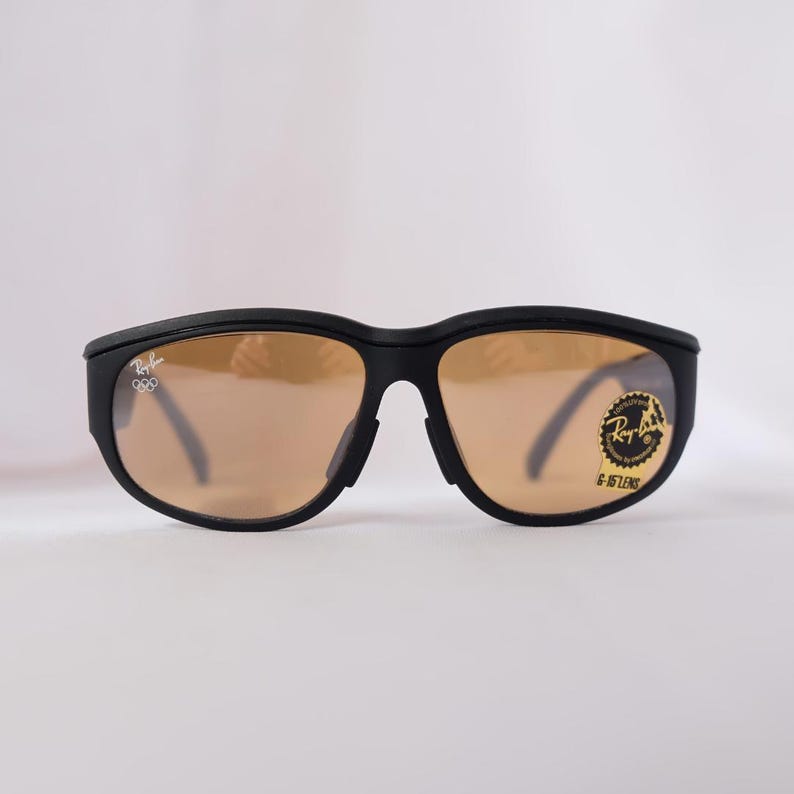 May include: Black wraparound sunglasses with brown lenses. The sunglasses have a Ray-Ban logo on the right lens and the text "G-15 LENS" on the right temple.