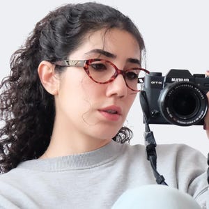 Op de afbeelding: Een vrouw met een bril met schildpadpatroon en een grijze sweatshirt houdt een zwarte Fujifilm digitale camera vast. De camera heeft een lens en een riem. De bril heeft een cat-eye vorm.
