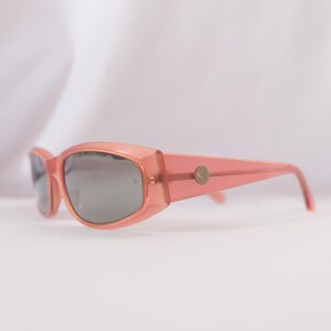 Pode incluir: Um par de &oacute;culos de sol cor-de-rosa com lentes cinzentas. Os &oacute;culos t&ecirc;m uma forma retangular com bordas arredondadas e um pequeno emblema dourado nas hastes. A arma&ccedil;&atilde;o &eacute; de cor s&oacute;lida e transl&uacute;cida.