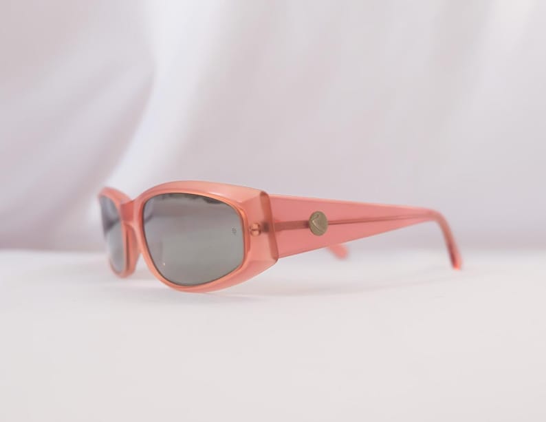 Pode incluir: Um par de &oacute;culos de sol cor-de-rosa com lentes cinzentas. Os &oacute;culos t&ecirc;m uma forma retangular com bordas arredondadas e um pequeno emblema dourado nas hastes. A arma&ccedil;&atilde;o &eacute; de cor s&oacute;lida e transl&uacute;cida.