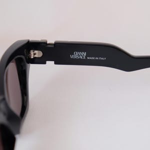 以下が含まれることがあります： 黒いサングラスのクローズアップ。レンズは濃い色です。「GIANNI VERSACE MADE IN ITALY」の文字がテンプルに印刷されています。サングラスは洗練されたモダンなデザインです。