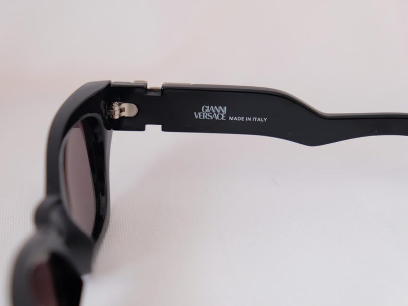 以下が含まれることがあります： 黒いサングラスのクローズアップ。レンズは濃い色です。「GIANNI VERSACE MADE IN ITALY」の文字がテンプルに印刷されています。サングラスは洗練されたモダンなデザインです。