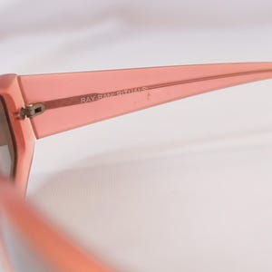 Pode incluir: Close-up de &oacute;culos de sol cor coral com o texto "RAY-BAN RITUALS" impresso na haste. Os &oacute;culos t&ecirc;m uma arma&ccedil;&atilde;o grossa e um design cl&aacute;ssico.
