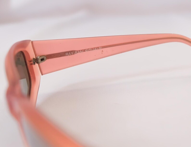 Pode incluir: Close-up de &oacute;culos de sol cor coral com o texto "RAY-BAN RITUALS" impresso na haste. Os &oacute;culos t&ecirc;m uma arma&ccedil;&atilde;o grossa e um design cl&aacute;ssico.