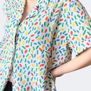 Puede incluir: Camisa blanca con estampado de confeti colorido. La camisa de manga corta tiene cuello y está decorada con formas rojas, amarillas, verdes y azules. La camisa está hecha de tela ligera.