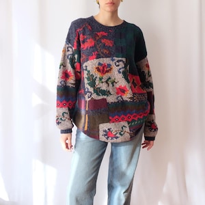 Op de afbeelding: Een patchwork trui met een ronde hals. De trui heeft bloemen- en geometrische patronen in rood, groen, blauw en bruin. De mouwen en de halslijn zijn effen marineblauw.