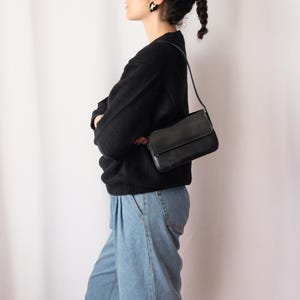 May include: A black leather shoulder bag with a rectangular shape and a flap closure. The bag is worn over the shoulder. The person is wearing a black sweater and light blue jeans.