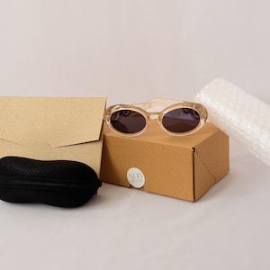 Pode incluir: &Oacute;culos de sol ovais com arma&ccedil;&atilde;o castanho-clara e lentes escuras sobre uma pequena caixa de cart&atilde;o. Uma capa preta para &oacute;culos de sol, um envelope castanho e um rolo de pl&aacute;stico bolha tamb&eacute;m est&atilde;o presentes.