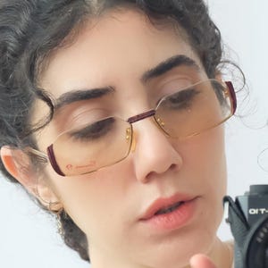 May include: Gold-rimmed eyeglasses with burgundy accents on the temples. The lenses are tinted, and the glasses have a rectangular shape. The image shows a close-up of the glasses on a person's face.