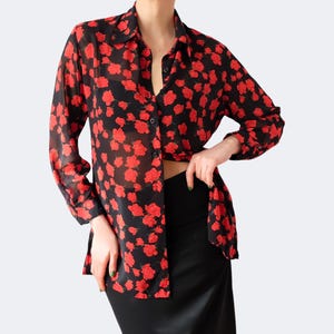 Pode incluir: Uma camisa preta com estampa floral vermelha. A camisa de manga comprida está parcialmente desabotoada. A modelo está usando uma saia preta. A camisa tem gola e botões na frente.