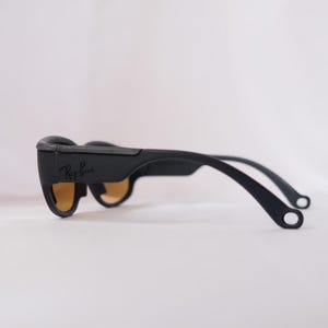May include: Black Ray-Ban sunglasses with brown lenses and a black frame. The sunglasses have a curved shape and a thin, flexible arm.