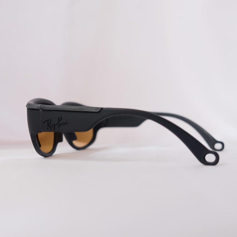 May include: Black Ray-Ban sunglasses with brown lenses and a black frame. The sunglasses have a curved shape and a thin, flexible arm.