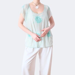 Op de afbeelding: Een lichtgroene top met een ronde hals en korte mouwen, versierd met turquoise bloemmotieven. Gecombineerd met een witte broek. Een ketting met hanger is ook zichtbaar. De outfit oogt luchtig.