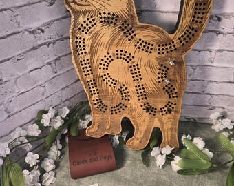 May include: A wooden cribbage board in the shape of a cat, featuring an engraved design and numerous peg holes. A small leather pouch labelled "Cards and Pegs" is present, along with playing cards and white flowers for decoration.