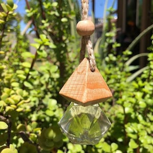 May include: A wooden pyramid-shaped car air freshener with a clear glass bottle filled with a green liquid. The air freshener is hanging from a string with a wooden bead.