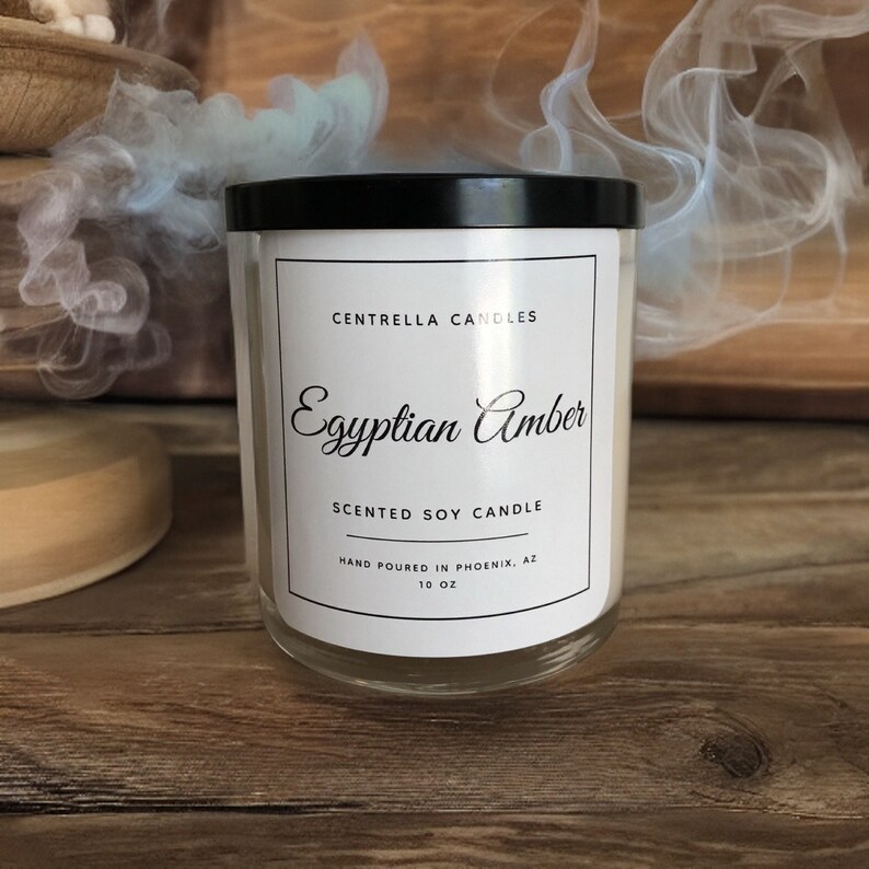 May include: A clear glass jar candle with a black lid. The label reads "Egyptian Amber Scented Soy Candle" from Centrella Candles, hand-poured in Phoenix, AZ. The candle is 10 oz. and sits on a wooden surface.