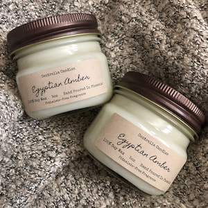 May include: Two small glass jar candles with brown lids. The labels read "Egyptian Amber" and "100% Soy Wax, 5oz, Hand Poured In Phoenix, Phthalate-Free Fragrance". The candles are on a textured, gray surface.
