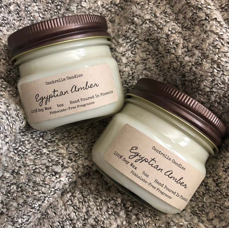 May include: Two small glass jar candles with brown lids. The labels read "Egyptian Amber" and "100% Soy Wax, 5oz, Hand Poured In Phoenix, Phthalate-Free Fragrance". The candles are on a textured, gray surface.