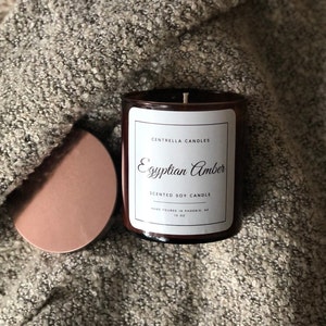 May include: A brown glass jar candle with a white label that reads "Egyptian Amber" and "Scented Soy Candle." The candle is 10 oz. and has a copper-colored lid. The candle sits on a gray, textured surface.