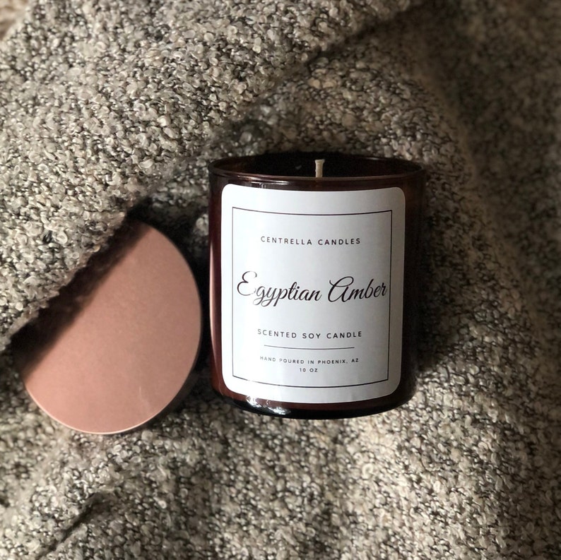May include: A brown glass jar candle with a white label that reads "Egyptian Amber" and "Scented Soy Candle." The candle is 10 oz. and has a copper-colored lid. The candle sits on a gray, textured surface.